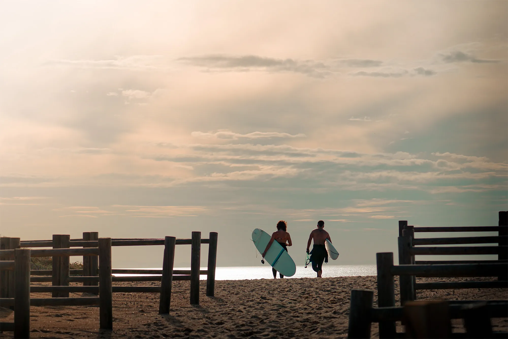 Dos surfistas caminando hacia el mar al atardecer entre vallas de madera